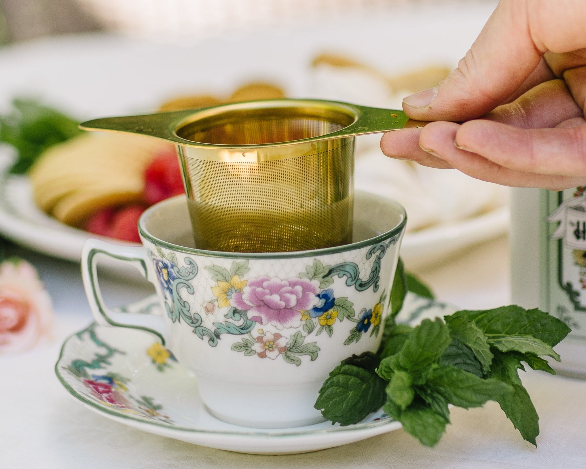 Metal tea strainer inside a teacup on a white tablecloth with a plate of food in the background.