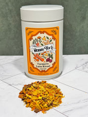 “Turmeric Latte Ritual tea tin with a pile of golden loose-leaf herbal blend on a white tabletop.”
