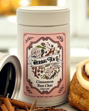Herbal Tea Co. cinnamon bun chai tea canister with cinnamon sticks and a cinnamon roll on a blurred background