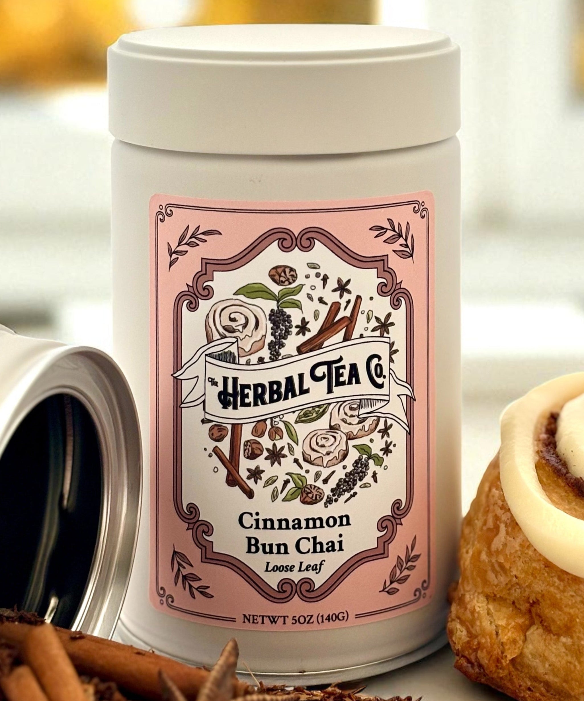 Herbal Tea Co. cinnamon bun chai tea canister with cinnamon sticks and a cinnamon roll on a blurred background