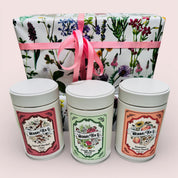 Three herbal tea tins with decorative labels in front of a floral gift box with a pink ribbon.