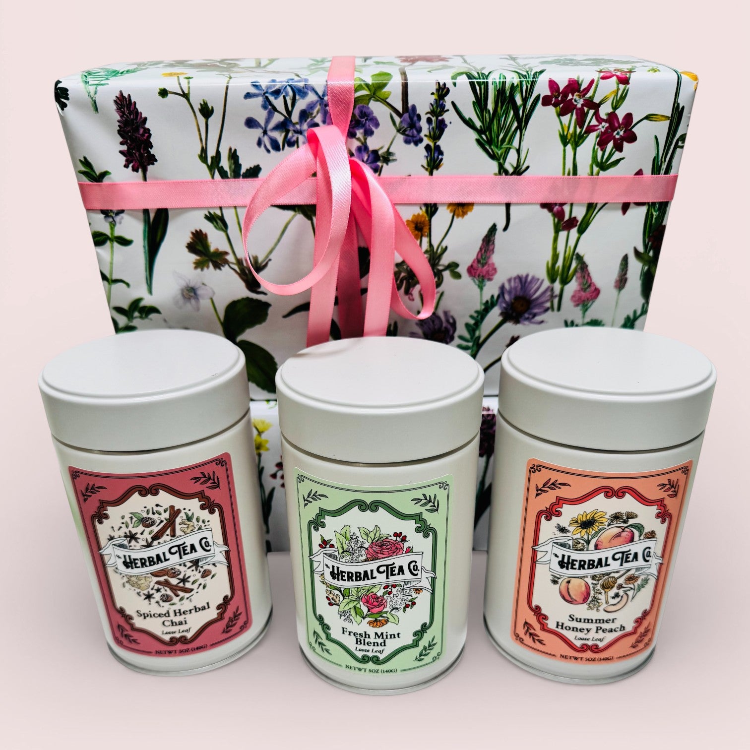 Three herbal tea tins with decorative labels in front of a floral gift box with a pink ribbon.